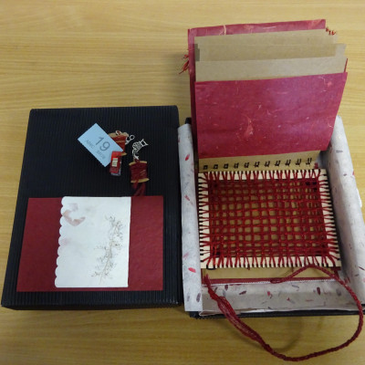 Small red weaving on a frame. Part of a ring-bound book, in an open box.